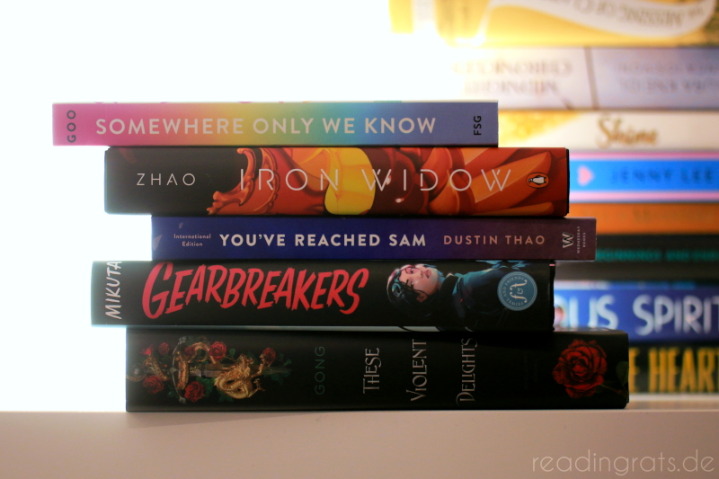Stack of five books in front of a light source on the left and another blurred out stack of books on the right. The stack is not neat but all backs face the camera. The books are from top down "Somewhere Only We Know", "Iron Widow", "You've Reached Sam", "Gearbreakers", and "These Violent Delights".